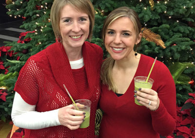 two people smile with their Pacific Perks green smoothies in front of a Christmas tree