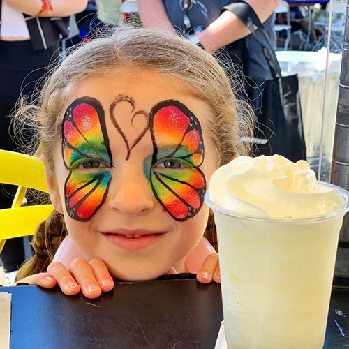 a Pacific Perks Italian soda on a table in front of a child with rainbow butterfly face paint